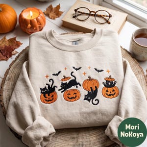 May include: A beige crewneck sweatshirt featuring an embroidered design of four black cats interacting with orange jack-o'-lanterns, accented by small orange stars and black bats. The embroidery is centered on the chest of the garment.