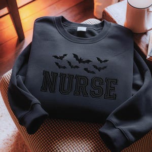 May include: A navy blue sweatshirt with the word "NURSE" in black block letters and a cluster of black bat silhouettes above it. The sweatshirt is folded and resting on a patterned chair.