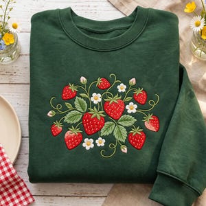 May include: A dark forest green crewneck sweatshirt featuring a central embroidered design of bright red strawberries, green leaves, and small white flowers with yellow centers. The embroidery is detailed with curved stems and vines.