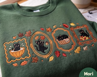 Op de afbeelding: Een donkergroene sweater met ronde hals voorzien van borduursels van zwarte katten in decoratieve kaders. De ontwerpen tonen katten in een kom, spelend met herfstbladeren en een eikel, slapend in een mandje en zittend in een kader.