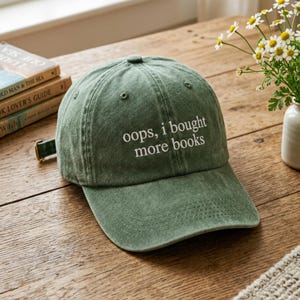 May include: A green baseball cap with the embroidered text "oops, i bought more books." The cap has a curved brim and an adjustable strap. The hat is sitting on a wooden surface.