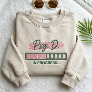 May include: A beige sweatshirt with the embroidered text "Psy. D." in green script over a pink brushstroke. Below, a green and pink loading bar reads "IN PROGRESS...". Sunglasses rest above the sweatshirt.