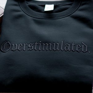 Puede incluir: Una sudadera negra con la palabra "Overstimulated" bordada en una fuente g&oacute;tica. La sudadera est&aacute; doblada y parece estar hecha de un material suave. El texto es de un tono negro m&aacute;s oscuro.