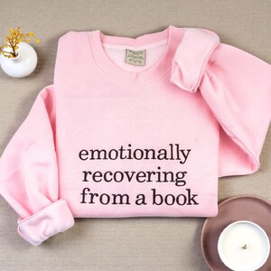 May include: A light pink long-sleeve crewneck sweatshirt featuring the black printed text "emotionally recovering from a book" centred on the chest. The garment has a ribbed collar, cuffs, and hem, with a visible interior brand label at the neck.