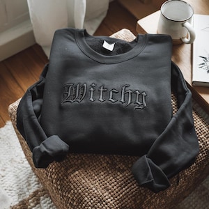 May include: A black long-sleeve sweatshirt featuring the word Witchy embroidered in a matching black thread across the chest in a gothic-style font. The garment has a crew neckline and long sleeves with ribbed cuffs, showcasing a solid dark color.