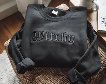 Op de afbeelding: Een zwart sweatshirt met lange mouwen en het woord Witchy geborduurd in bijpassende zwarte draad op de borst in een gotisch lettertype. Het kledingstuk heeft een ronde hals en lange mouwen met geribbelde boorden in een effen kleur.