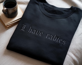 May include: A black crewneck sweatshirt featuring the text I have rabies embroidered in a dark, gothic-style font across the chest. The garment is made of a soft, fleece-lined fabric and is presented folded neatly on a textured, light-colored surface.