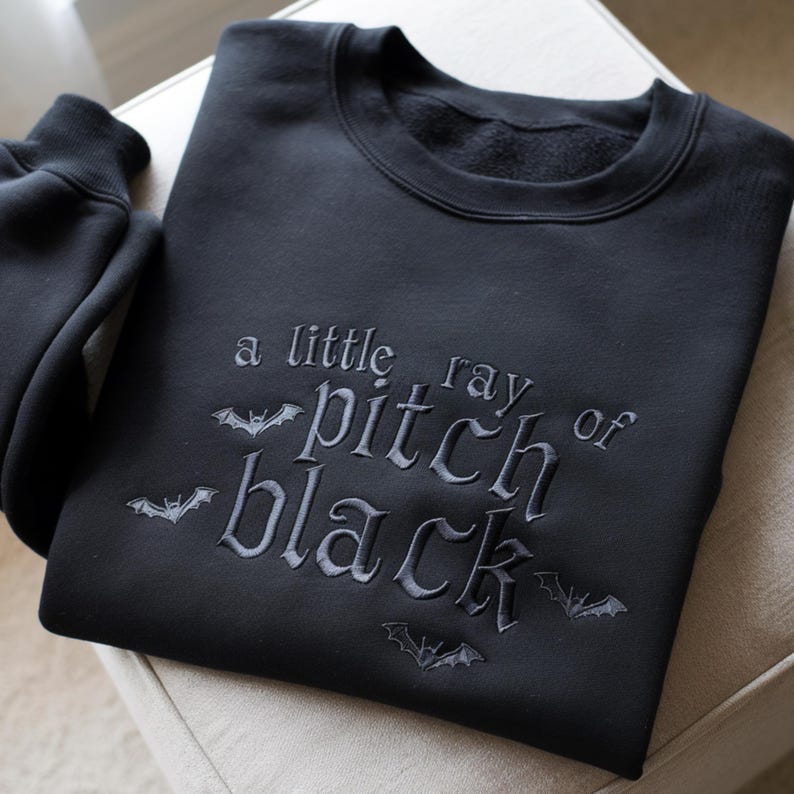 May include: Black sweatshirt with the embroidered phrase 'a little ray of pitch black' in a gothic font. The phrase is accompanied by bat illustrations. The sweatshirt is made of a soft material and is displayed on a neutral surface.