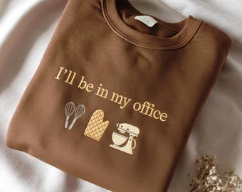 May include: A brown crewneck sweatshirt featuring the embroidered text "I'll be in my office" in a light yellow thread. Below the text are embroidered icons of two whisks, a quilted oven mitt, and a stand mixer in shades of yellow and white.