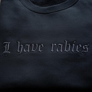 May include: Black sweatshirt with the embroidered phrase "I have rabies" in a gothic font. The text is in a lighter shade of black than the sweatshirt. The sweatshirt has a crew neck.