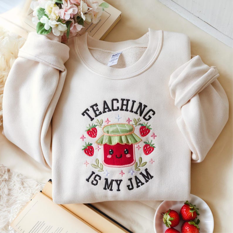 May include: Cream-colored sweatshirt with the embroidered phrase "TEACHING IS MY JAM." The design features a smiling jar of jam surrounded by strawberries and leaves. The sweatshirt has long sleeves with rolled cuffs.