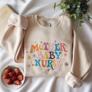 May include: A cream-colored crewneck sweatshirt featuring colorful embroidered text that reads MOTHER BABY NURSE in a wavy font. The design is accented with small embroidered stars and hearts in shades of pink, blue, orange, and yellow.