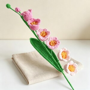 Könnte beinhalten: Ein handgefertigter Häkelblumenstiel mit sechs rosa glockenförmigen Blüten und leuchtend gelben Zentren. Der Stiel ist leuchtend grün und hat ein langes, strukturiertes Blatt an der Basis, das aufwendige Garnarbeit und detaillierte Stiche zeigt.