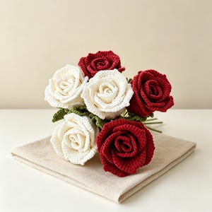 Könnte beinhalten: Ein Strauß aus fünf gehäkelten Rosen mit drei dunkelroten und zwei cremefarbenen Blüten. Jede Rose ist aus detaillierten Garnblütenblättern gefertigt und wird von kleinen, strukturierten grünen gehäkelten Blättern an der Basis ergänzt.
