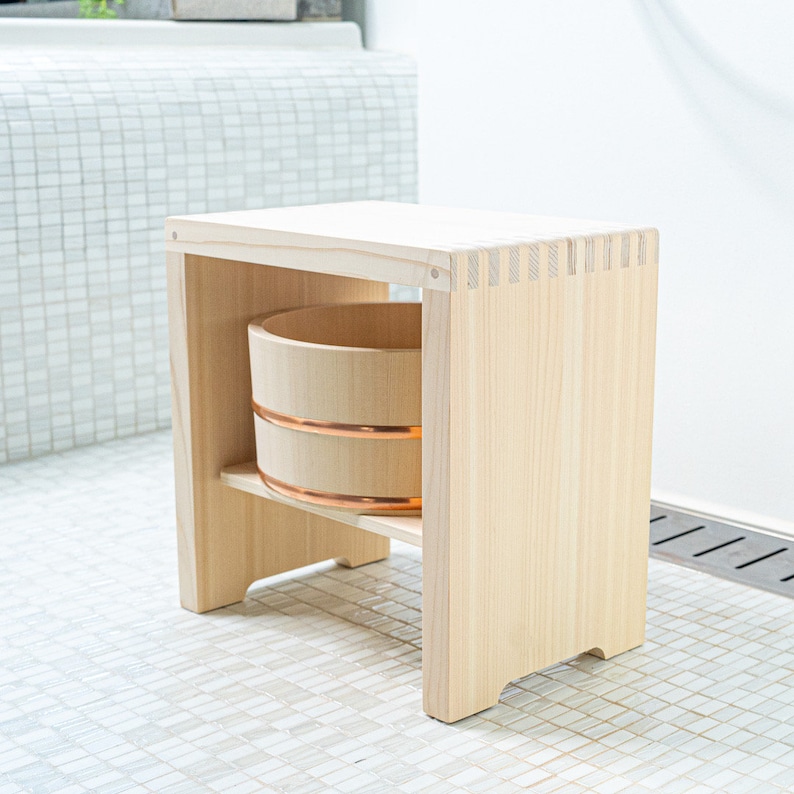 Minimalist Japanese wooden bath stool with a natural finish, featuring a neatly stacked hinoki wood bucket beneath, placed in a serene bathroom setting with greenery in the background.
