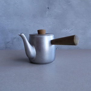 Può includere: Una teiera d'argento con un manico di legno e un coperchio di legno. La teiera è su una superficie grigia.