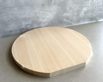 May include: A circular wooden cutting board with a light beige color and a visible straight grain pattern. The board has a smooth, flat surface and a thick, rounded edge. It is partially wrapped in a thin, clear plastic film for protection.
