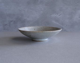 May include: A handmade, light gray ceramic bowl with a wide, shallow design. The bowl has a slightly textured surface and a small, rounded base. The interior and exterior are a muted gray color with subtle variations. The bowl is suitable for serving or display.