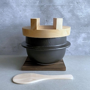 May include: A black ceramic rice pot with a wooden lid and handle, sitting on a dark wooden base. A wooden rice paddle rests in front. The pot has a rounded shape and a light-colored wooden lid.