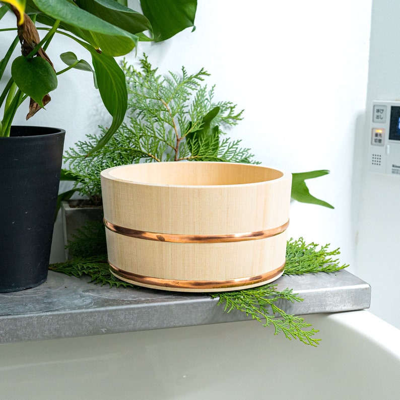 Traditional Japanese hinoki wood bath bucket with copper bands, elegantly placed on a stone ledge surrounded by lush greenery, evoking an authentic onsen experience.