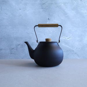 Puede incluir: Una tetera negra mate con un asa de madera y una tapa de corcho. La tetera tiene un cuerpo redondeado y un pico curvo. El asa es de metal y madera. El fondo es una pared azul claro.