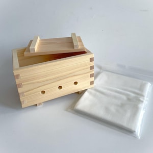 May include: A wooden tofu press with a lid and a white cloth. The press has a rectangular shape with a series of holes on the bottom. The cloth is folded and placed in a plastic bag.