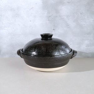 May include: A round, black ceramic cooking pot with a matching lid and two side handles. The pot features a glossy, dark finish on the upper portion and lid, while the base remains a contrasting, unglazed light beige color with a smooth texture.
