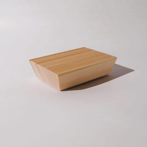 Puede incluir: Una jabonera rectangular de madera clara que presenta una serie de ranuras horizontales paralelas en la superficie superior. El objeto tiene lados inclinados y un acabado de veta de madera natural, presentado desde una perspectiva de ángulo alto.
