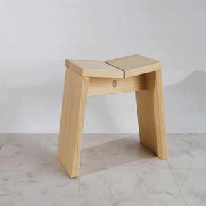 Puede incluir: Un taburete de madera de color claro con un diseño de asiento dividido e inclinado y dos patas robustas y acampanadas. La viga de soporte frontal muestra un pequeño logotipo circular estampado en la veta de la madera, con acabado natural.
