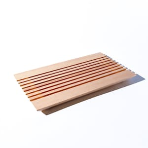 May include: A light-colored wooden soap dish with a slatted design. The rectangular dish has parallel grooves across its surface, allowing for drainage. The wood has a natural grain and a warm, inviting tone. The dish is designed to hold a bar of soap.