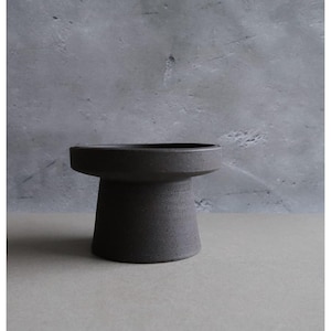 Peut inclure: Un bol sur piédestal en céramique gris foncé, avec un dessus large et plat et une base cylindrique. Le bol a une surface texturée et un design minimaliste, adapté pour exposer de petits objets ou comme élément décoratif. Le bol mesure environ 15 cm de diamètre.
