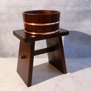 May include: A dark brown wooden Japanese soaking tub with copper bands sits on a matching wooden stool. The tub has a cylindrical shape and is made of vertical wooden slats. The stool has a simple, angular design.