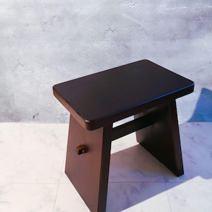 Könnte beinhalten: Ein dunkelbrauner Holzstuhl mit rechteckiger Sitzfläche und abgewinkelten Beinen. Der Hocker hat einen kleinen, runden Holzstift an der Seite. Der Hocker ist etwa 46 cm lang, 30 cm breit und 41 cm hoch.