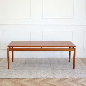 Op de afbeelding: Een moderne teakhouten salontafel uit het midden van de eeuw met een rechthoekig blad en een eenvoudig, minimalistisch ontwerp. De tafel heeft een lichtbruine afwerking en wordt ondersteund door vier slanke poten.