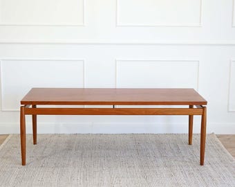 May include: A mid-century modern teak coffee table with a rectangular top and a simple, minimalist design. The table has a light brown finish and is supported by four slender legs.