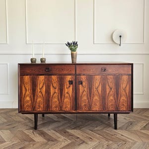 Op de afbeelding: Een rechthoekig houten dressoir met een rijke, donkerbruine houtnerf, voorzien van twee bovenlades en twee onderste kastdeuren. Het meubelstuk rust op vier taps toelopende poten en heeft geïntegreerde vierkante houten handgrepen op de lades en deuren.
