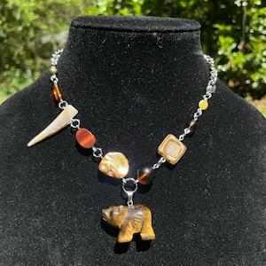 May include: A necklace with a silver chain and a variety of beads in shades of brown, amber, and gold. The necklace features a carved bear pendant and a horn-shaped charm. The beads include round, square, and irregular shapes.