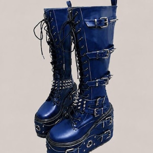 May include: A pair of navy blue platform boots with multiple buckles, silver-tone studs, and lace-up closures. The boots have a high shaft and a chunky platform sole, creating a bold, edgy aesthetic. The boots are made of a shiny material.