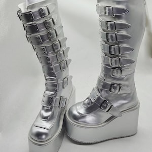 May include: A pair of silver platform boots. The boots have multiple straps with buckles and a thick platform sole. The boots are knee-high and made of a shiny, metallic material. The buckles and hardware are also silver.