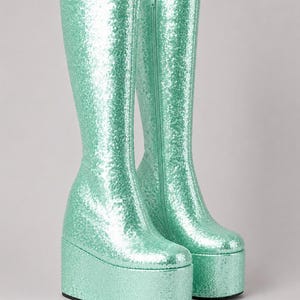 Könnte beinhalten: Ein Paar mintgrüne Plateau-Stiefel. Die kniehohen Stiefel haben eine klobige Plateausohle und einen seitlichen Reißverschluss. Der gesamte Stiefel ist mit schimmernden Pailletten bedeckt, die einen funkelnden Effekt erzeugen. Retro-Design im Disco-Stil.