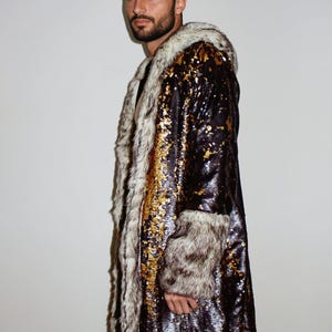 Puede incluir: Una larga túnica negra de lentejuelas con detalles dorados y ribete de piel sintética blanca. La túnica tiene capucha y mangas largas.