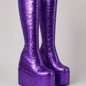Pode incluir: Um par de botas de plataforma até o joelho cobertas por lantejoulas roxas brilhantes. As botas têm uma sola de plataforma grossa e um fecho de correr na parte interna. As botas são uma declaração de moda vibrante.
