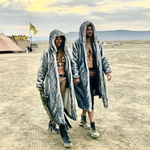 May include: Two people wearing grey faux fur hooded coats with black trim. The coats are long and have a shaggy texture. The people are standing in a desert landscape.
