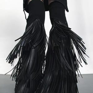 May include: Black knee-high boots with long, flowing fringe. The boots are made of a dark material and feature a sleek, modern design. The fringe adds a dramatic flair to the footwear, creating a bold and stylish look.