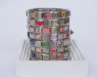 Op de afbeelding: Een stapel modulaire schakelarmbanden van roestvrij staal met diverse kleurrijke emaille bedels, waaronder een rode aardbei, een roze ster, een blauw huisje, een roze hartje, een paarse bloem en diverse geometrische patronen.