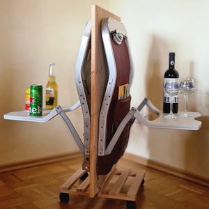 Op de afbeelding: Een opvouwbare serveerwagen met een houten frame, bruine lederen details en twee witte trays. De wagen heeft een houten basis met wielen en bevat een fles wijn, twee glazen en twee bierblikjes.
