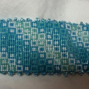 May include: A blue and white beaded bracelet with a geometric pattern. The bracelet is made of small, square beads and has a silver clasp.