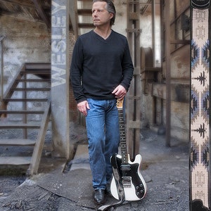 May include: A man standing in a rustic setting, holding a black and white electric guitar with a brown guitar strap. The guitar strap has a geometric pattern in shades of blue, brown, and white.
