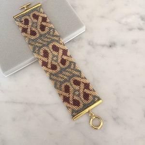 May include: A gold beaded bracelet with a geometric pattern in shades of grey, beige, and burgundy. The bracelet has a gold clasp and a toggle closure.