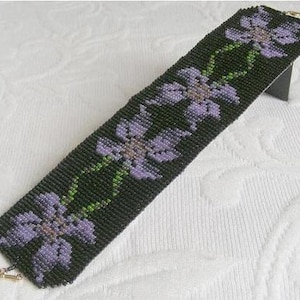 May include: A beaded bracelet featuring a black background and a pattern of purple flowers. The bracelet is crafted with small beads and has a delicate design.